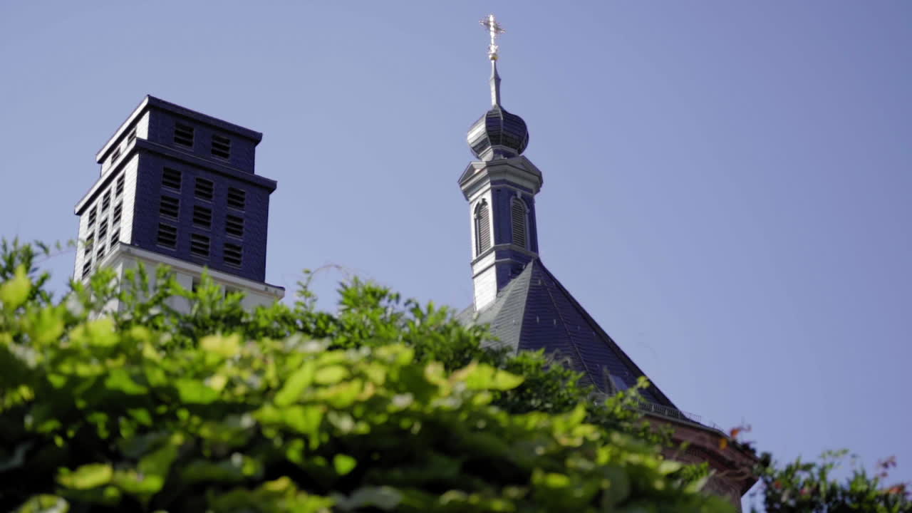 Slow-motion close-up of the Orangery Church tower in Blieskastel, Germany, framed by vibrant summer greenery and a clear blue sky. A stunning blend of nature and traditional architecture.