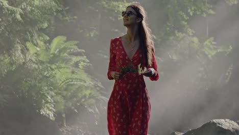 Enchanted-Forest-Stroll:-Close-up-of-a-woman-in-a-vibrant-red-dress-holding-a-juniper-branch,-walking-through-a-lush,-fairy-tale-forest-along-a-dried-riverbed-amid-the-mystical-morning-mist-and-fog