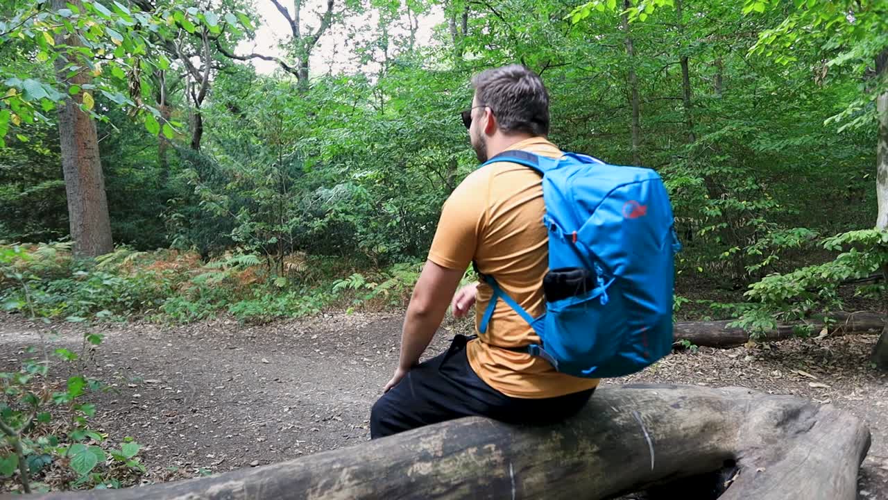 Tired and exhausted tourist sitting down on a fallen tree in the forest to take a break and relax. View from behind the back