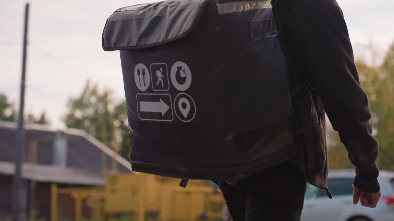 White male courier walking suburban street carrying large insulated delivery backpack, autumn foliage and residential houses in background, focused expression while checking smartphone for order