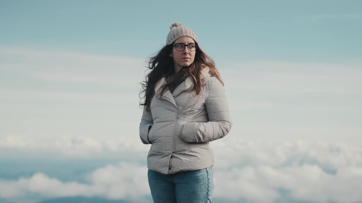 Una bella giovane donna con una giacca grigia e occhiali contempla la bellezza della natura ammirando le mozzafiato viste panoramiche delle montagne e delle nuvole.
