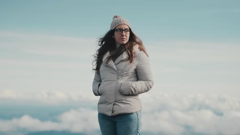 a beautiful young woman in a gray jacket and glasses contemplates the beauty of nature while taking in the stunning panoramic views of the mountains and clouds