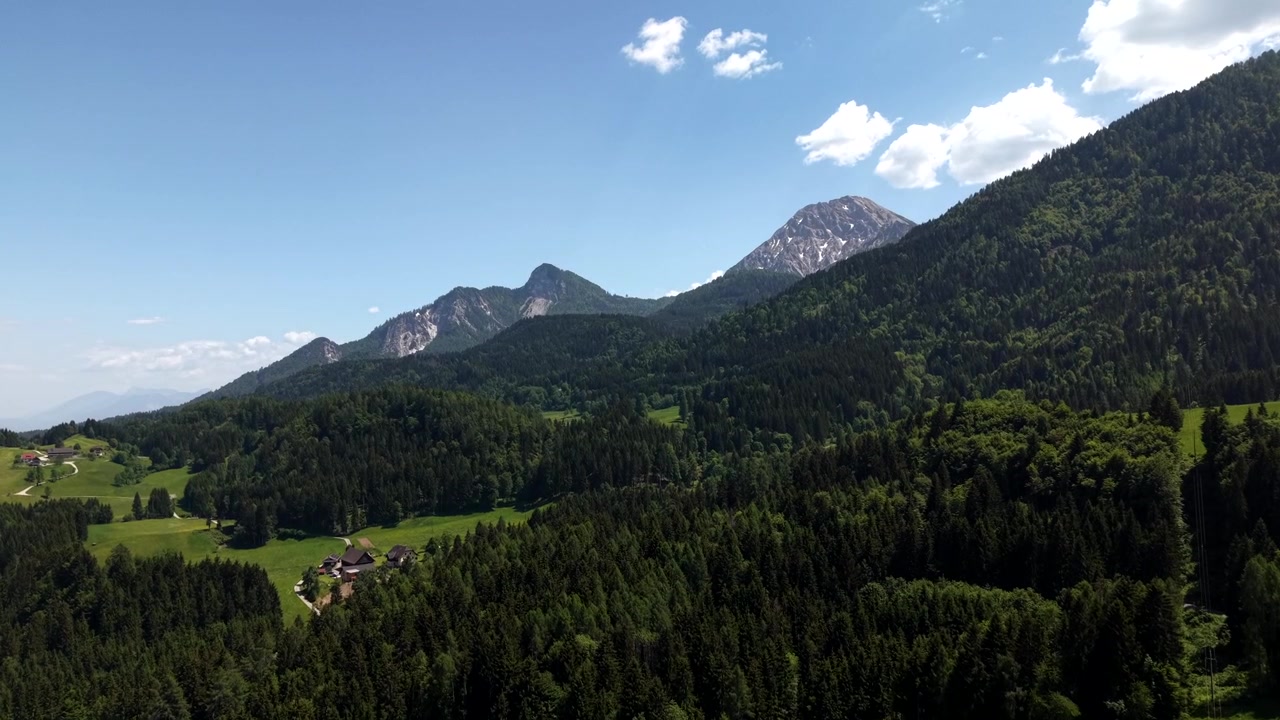 Rugged snow-streaked alpine peaks rise above dense forested hillsides in Austrian countryside, small farmhouse chalet sits beside green meadow beneath sunlit summer sky. Aerial drone rising wide