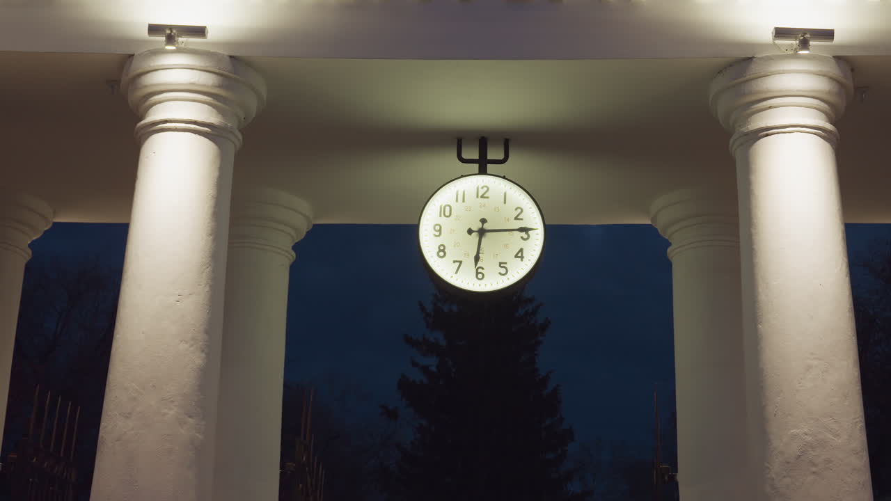 Large round illuminated clock positioned between four classic white columns, glowing against dark blue evening sky with distant silhouette of tree and moody architectural ambiance