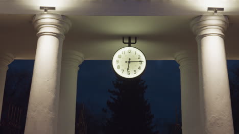 large round illuminated clock positioned between four classic white columns, glowing against dark blue evening sky with distant silhouette of tree and moody architectural ambiance