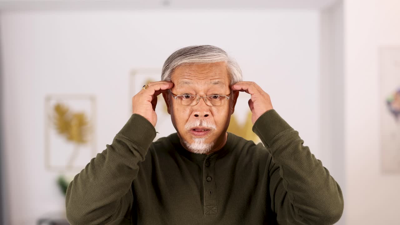 Senior Asian man sits indoors, pressing fingers to his temples with a pained expression, suggesting headache discomfort. Soft, natural lighting and steady camera enhance clarity