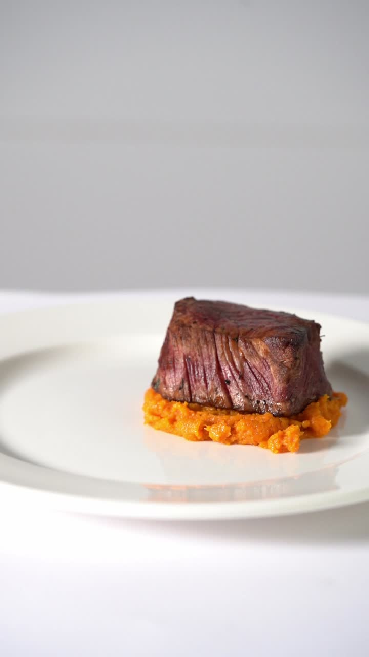 Succulenta bistecca di manzo alla griglia servita su purè di verdure cremoso in un piatto minimalista, primo piano.