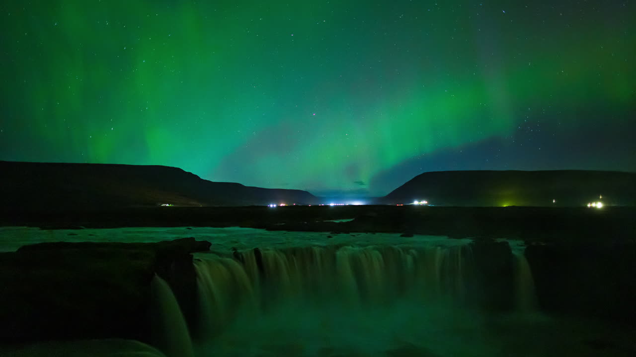 Aurora boreale alla cascata di Godafoss, Islanda. Cieli verdi drammatici e luminosi con luci del traffico e cascata in primo piano. Time lapse