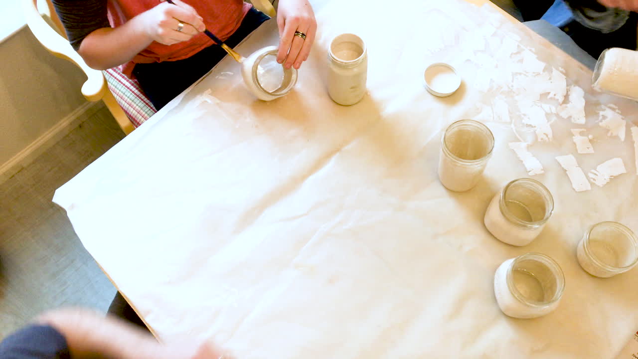 Top view of table at home with three people sitting at the table and painting glass jars with paint for wedding decoration