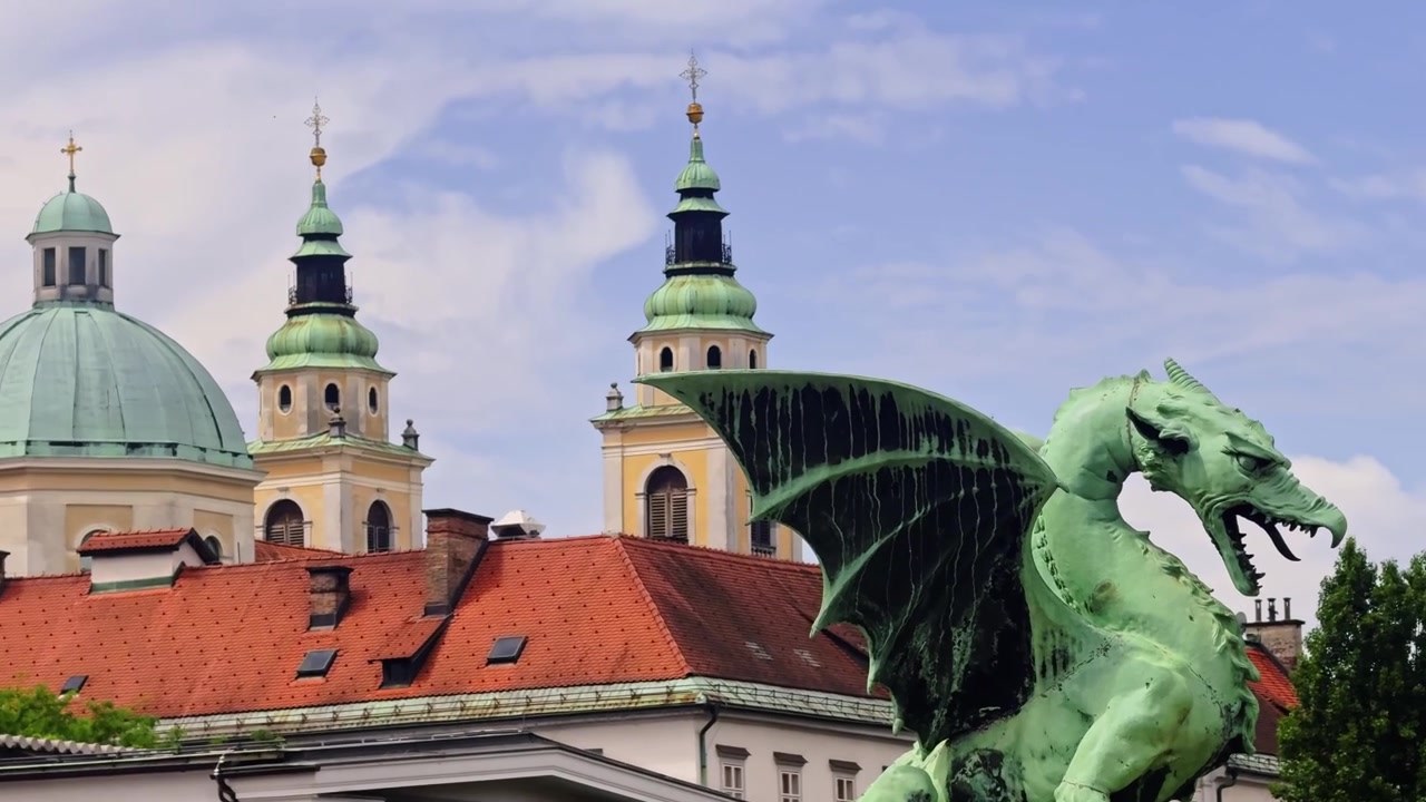 Statua del drago sul Ponte dei Draghi di Lubiana, con la Cattedrale di Lubiana, le torri gemelle della Cattedrale di San Nicola e i tetti rossi del centro storico sullo sfondo.