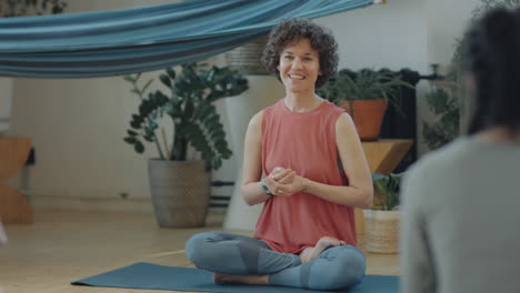 a woman smiles while sitting in a yoga pose on a mat in a bright studio during a class