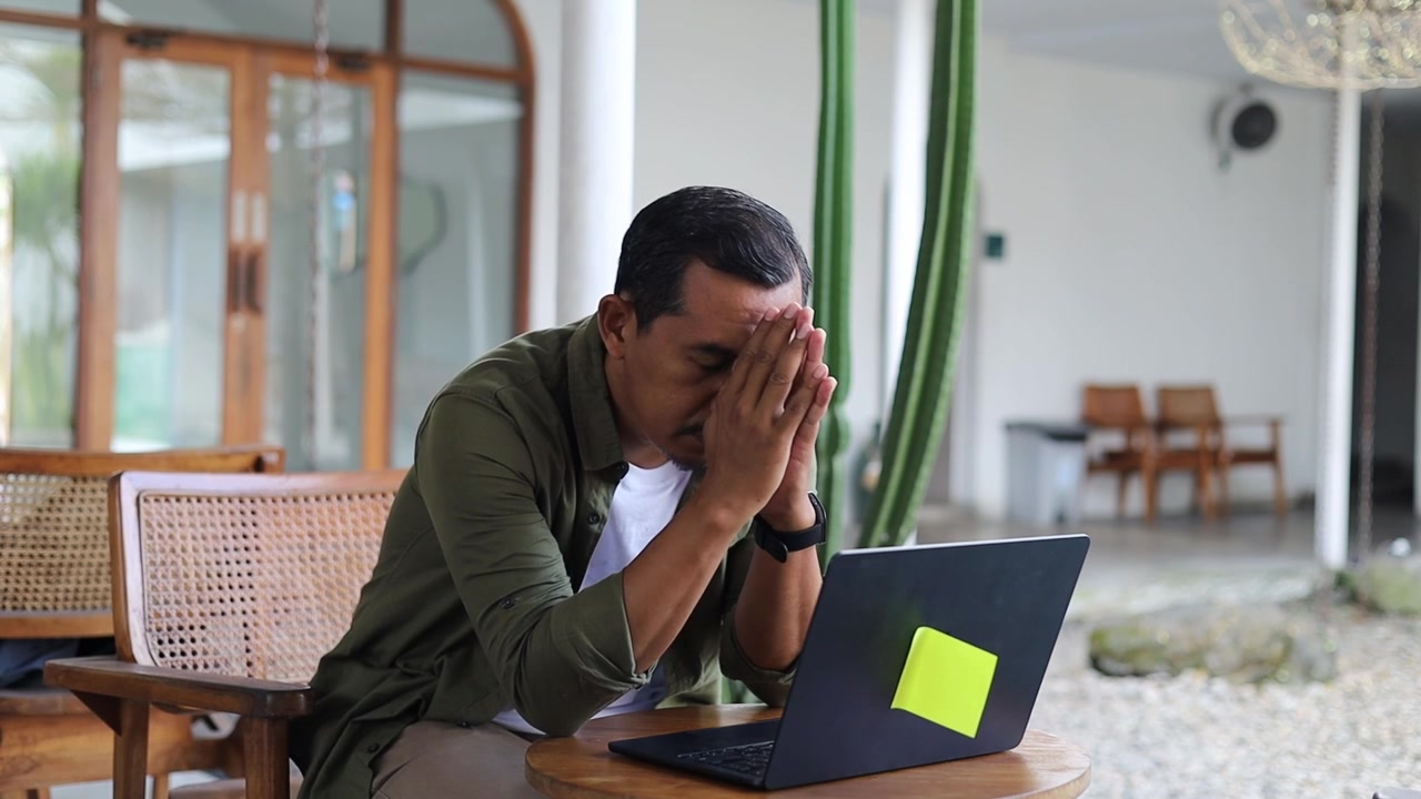 Overworked concentrated Asian copy-writer wearing shirt working on new articles using laptop computer, leaning his elbow on cafe table