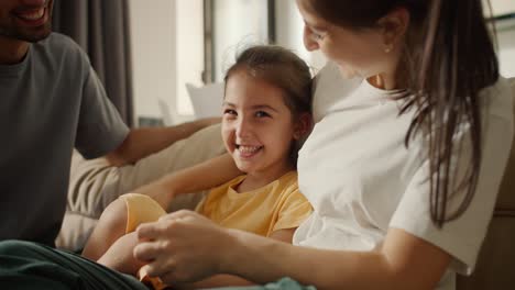 Close-up-shot-of-a-brunette-mom-in-a-white-t-shirt-playing-with-her-daughter-in-a-yellow-dress-while-sitting-on-a-light-brown-sofa-with-her-dad-in-a-modern-studio-room