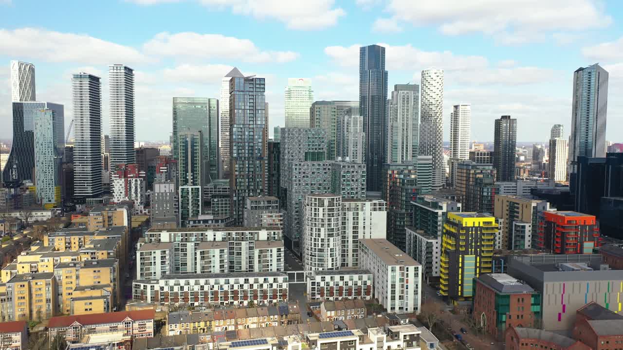 Aerial drone footage showing modern skyline of Canary Wharf in London, England, featuring glass skyscrapers, colorful buildings, and vibrant urban architecture under bright cloudy sky