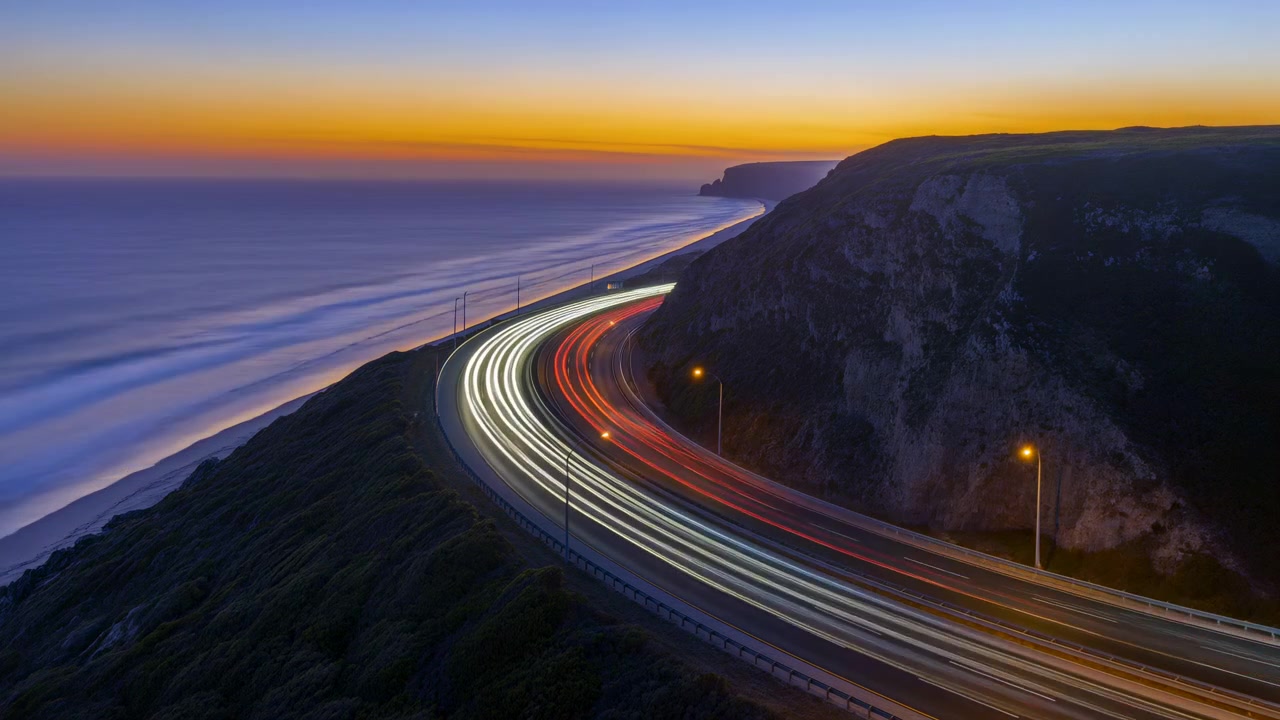 Vista a lunga esposizione mostra luci di veicoli bianche e rosse in movimento che seguono una strada costiera curva accanto a scogliere e oceano calmo durante la luce serale, con carreggiata illuminata e ampia vista sull'orizzonte.