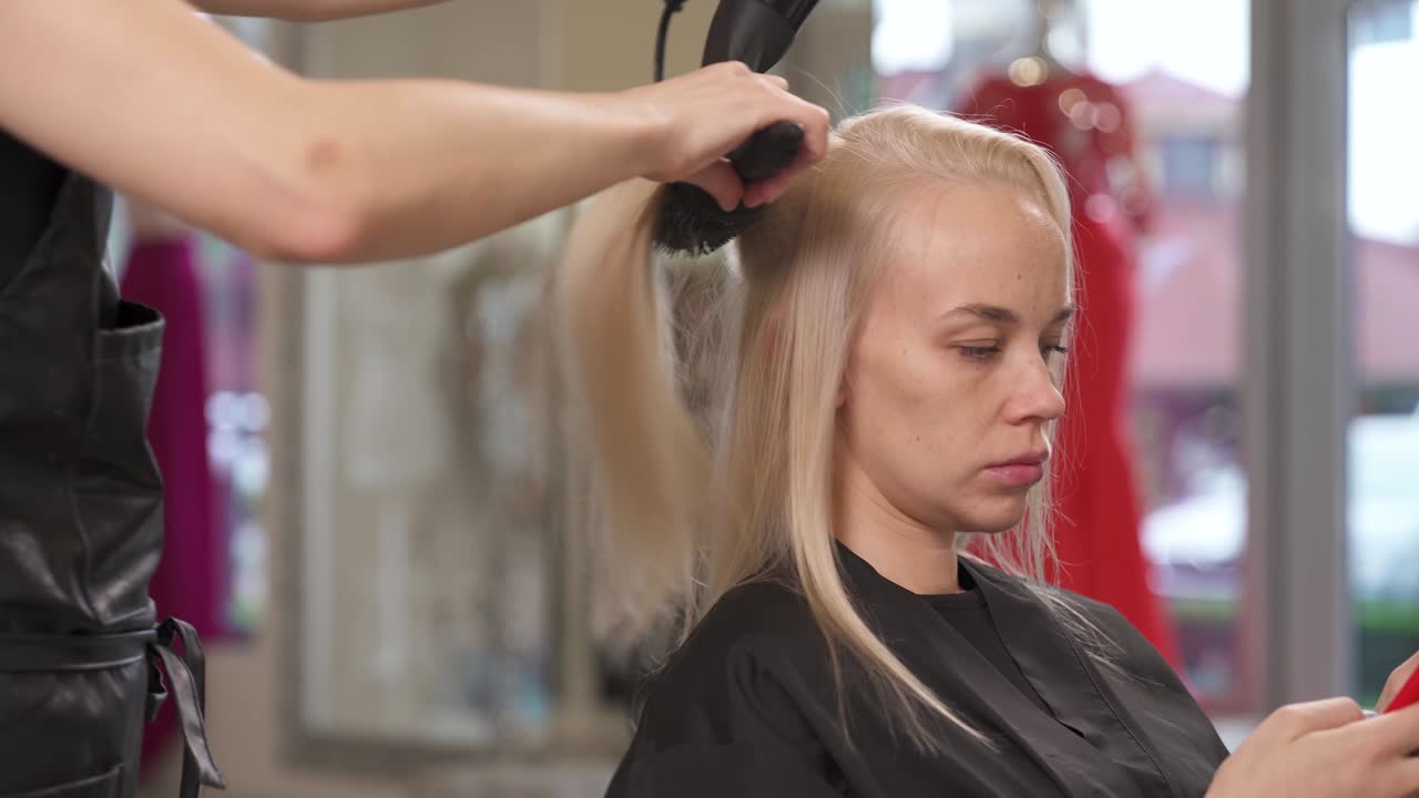 una vista lateral de la rubia sentada en la silla y escribiendo en el teléfono. la peluquera recoge el cabello en hebras con un pincel, lo estira y se seca con un secador de cabello.