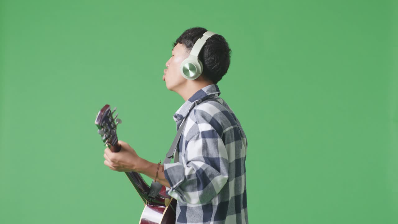 녹색 스크린 배경에서 기타를 연주하고 노래하는 헤드폰을 착용한 젊은 아시아 십대 소년의 측면 시각