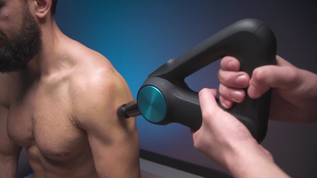 masajeador de percusión de primer plano en las manos de un masajista hace un masaje de los hombros y la parte superior de la espalda a un cliente masculino acostado