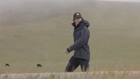 Adult-male-walks-along-highway-with-cows-grazing-fog-covered-hillside-in-background