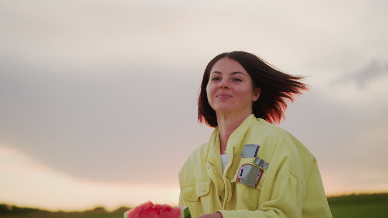 cata de sandía en un campo al atardecer disfrutando de la brisa de la pradera, mujer levantando una jugosa rodaja, riendo y dando un mordisco divertido, manos pegajosas, avispa captando la luz del borde, movimiento espontáneo, accesorios de picnic