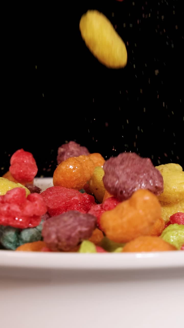 Pedaços de cereal vibrantes caem em cascata numa tigela branca contra um fundo escuro, criando uma cena dinâmica e colorida.