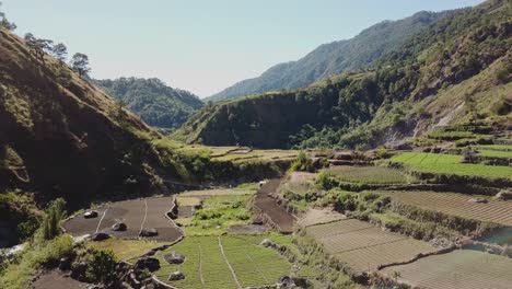 green-vegetable-garden-paddy-farms-in-mountainous-valley-village-in-Kabayan-Benguet-Philippines-wide-aerial-panning-right-to-left-revealing-river-blue-sky-bright-beautiful-sunny-day