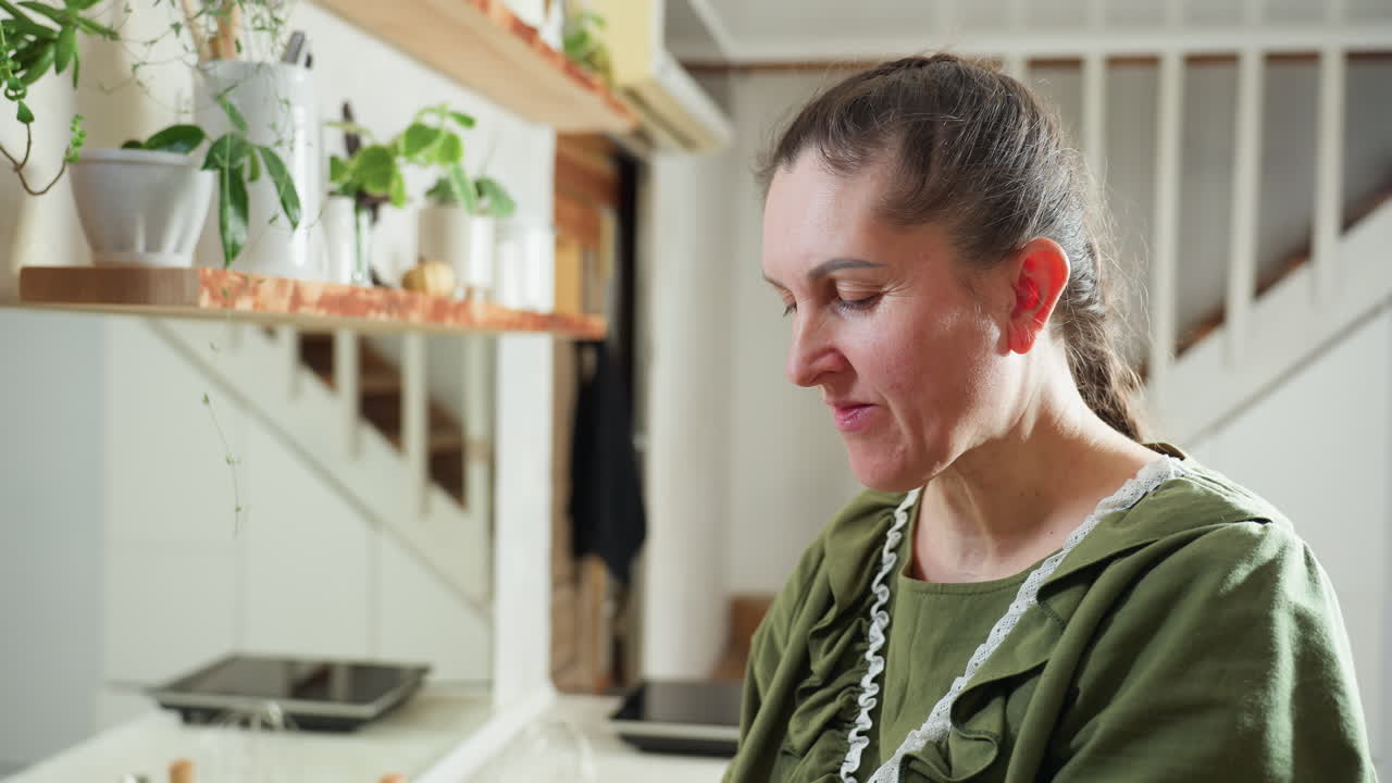 Middle-aged woman in green outfit joyfully eats fresh berry in bright kitchen while shaking head in satisfaction, expressing happiness and delight, surrounded by indoor plants