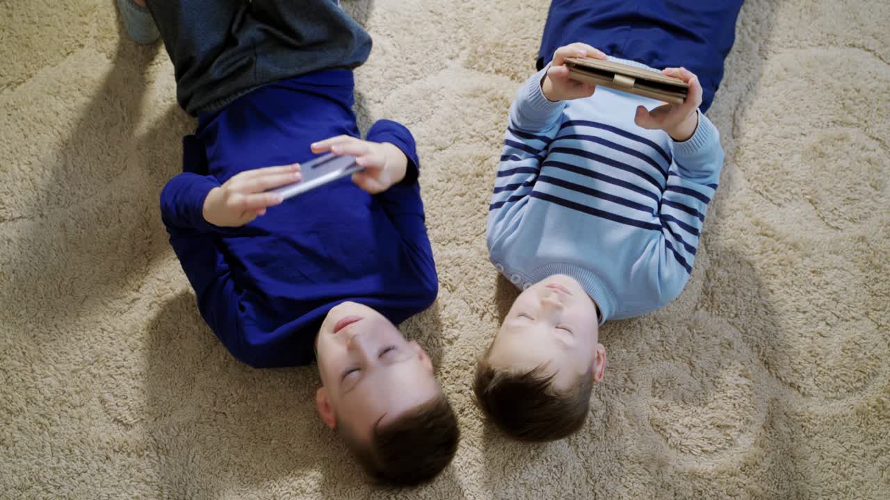 Boys lying on the floor on a light carpet and playing games on mobile phones at home. Top view of two kids playing together and helping each other in games.
