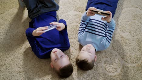 boys lying on the floor on a light carpet and playing games on mobile phones at home. top view of two kids playing together and helping each other in games.