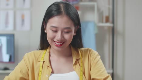 미소 짓는 아시아 여성 디자이너가 스튜디오에서 옷을 디자인하는 것을 즐기고 있습니다.