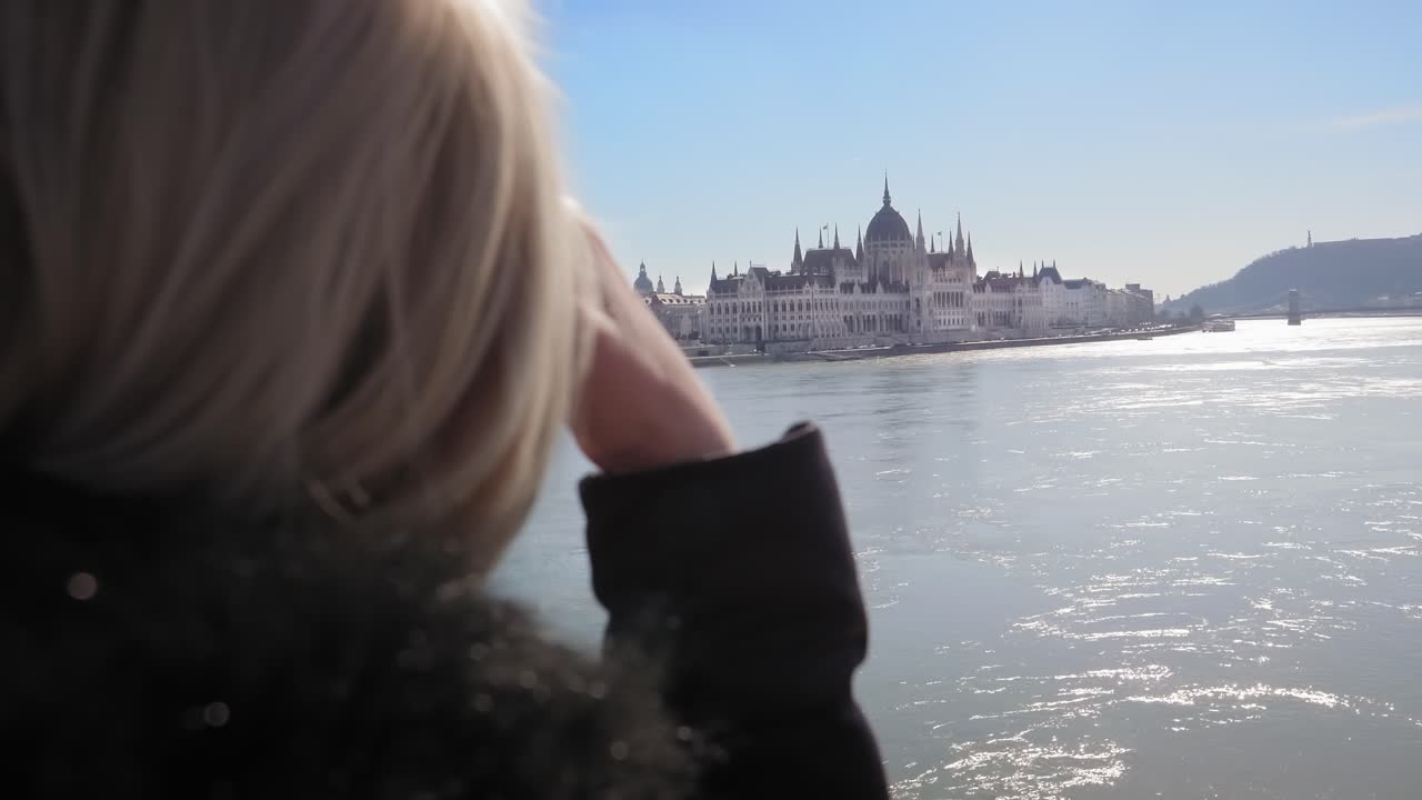 Young blonde woman admiring the view from the Margaret Bridge. Wide Danube river reflecting the sunlight. Hungarian Parliament in the background