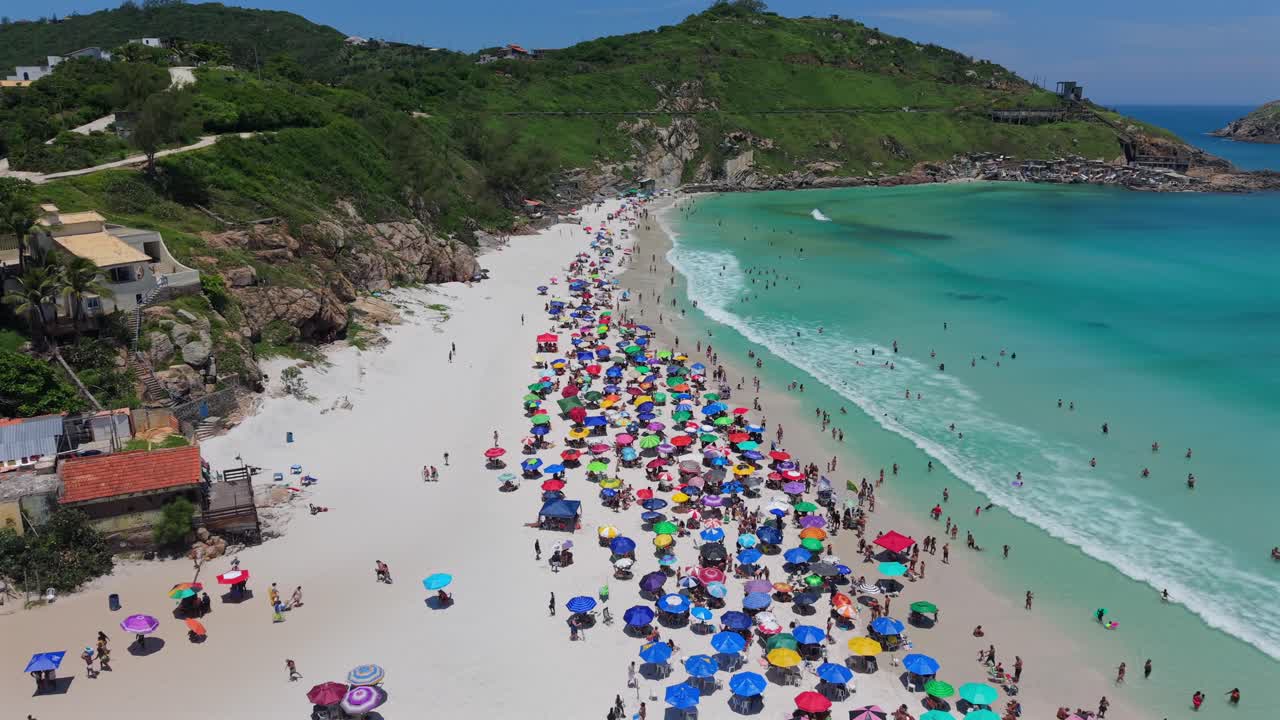 プライア・グランデのビーチパラソル。ターコイズブルーの海、大勢の人々、澄み切った空の下の緑の丘。ローアングルからの前方空中撮影。