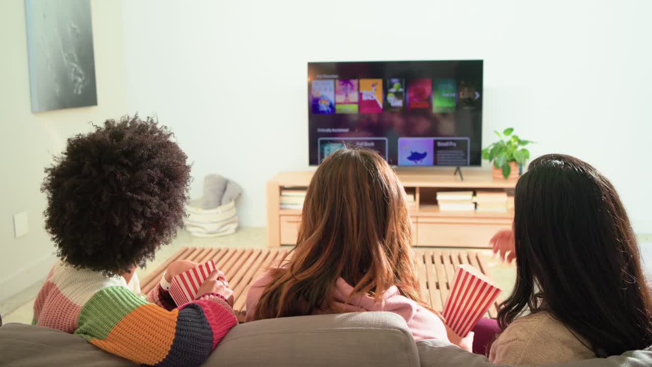 Amiche diverse guardano un menu in streaming sul divano, prendono popcorn e selezionano un titolo da guardare