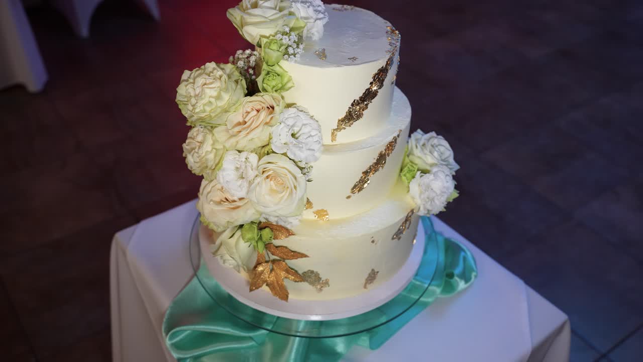 Close-up shot of a three-tiered wedding cake adorned with white and green floral decorations. Delicate lighting highlights the cake's intricate details, perfect for elegant celebrations