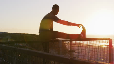 Fokussierter-Afroamerikanischer-Mann,-Der-Sich-Dehnt-Und-Bei-Sonnenuntergang-Im-Freien-Am-Meer-Trainiert