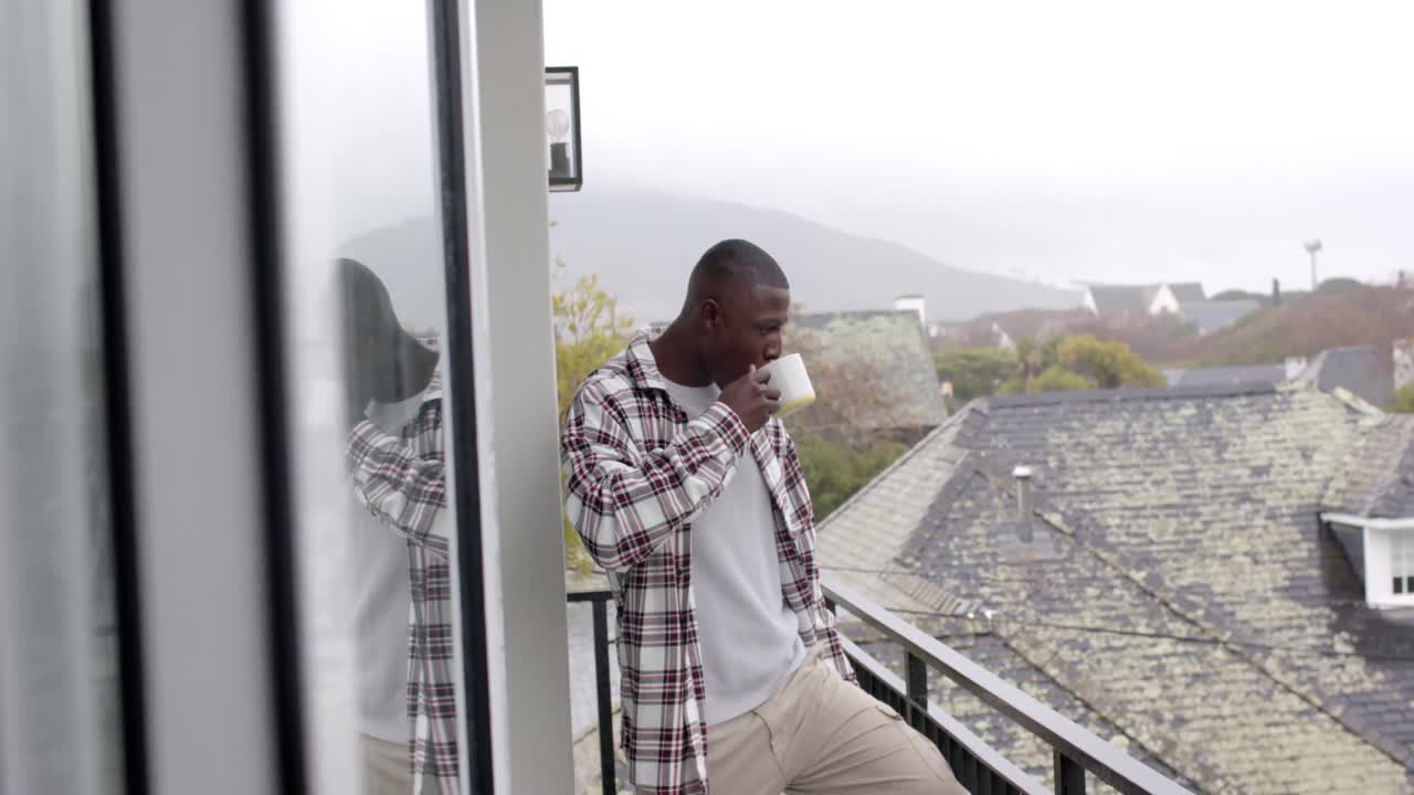 hombre afroamericano de pie en el balcón y bebiendo café en casa, cámara lenta