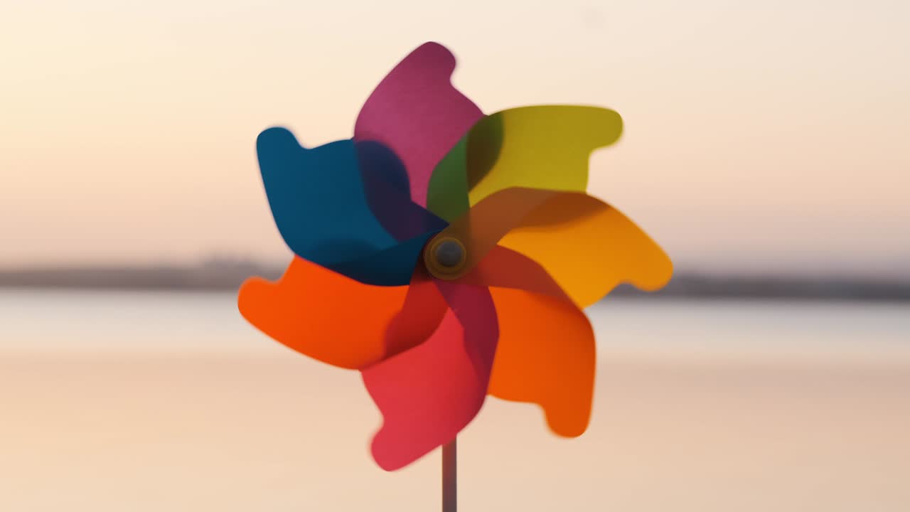 바다의 부드러운 표면과 밝은 분홍색 해가 지는 느린 동작에 대항하여 바다에서 모래에 서있는 바람을 불어넣는 플라스틱 핀을 회전하십시오. 해변에 장난감 밀. 태양의 큰 디스크.