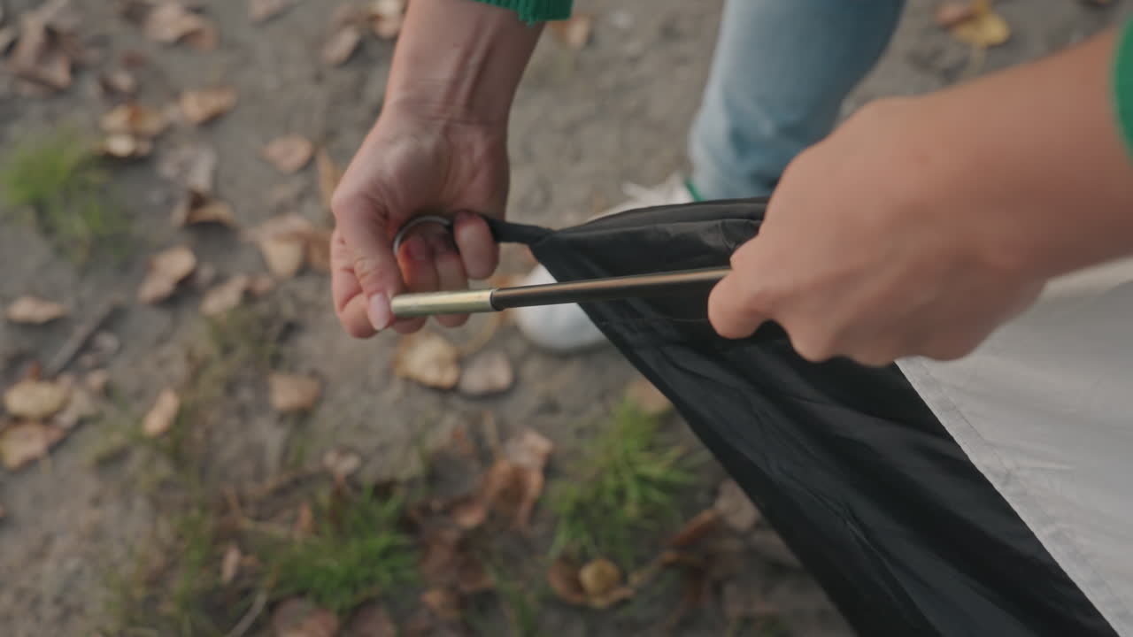 Campista asegurando la esquina de la tienda de campaña, manos insertando el extremo del poste en el ojal y la funda de tela, suelo cubierto de hojas de otoño, zapatillas blancas y vaqueros visibles, tomas de cerca del tensado.