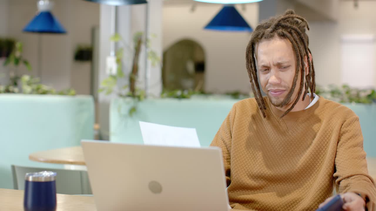Uomo africano che osserva attentamente un foglio stampato al tavolo di un caffè, consultando telefono e laptop mentre lavora.