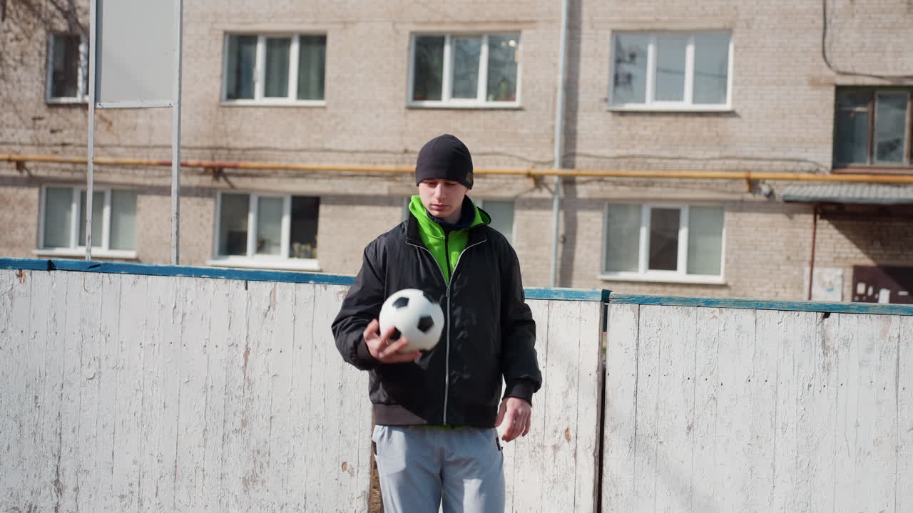 울타리 옆에서 축구공을 들고 친구와 마주보고 있는 흰색 티셔츠를 입은 10대 소년, 후드 재킷과 비니, 캐주얼 청바지, 아파트 단지 배경, 장난기 넘치는 긴장감과 움직임, 솔직한 도시 스포츠 순간, 패스