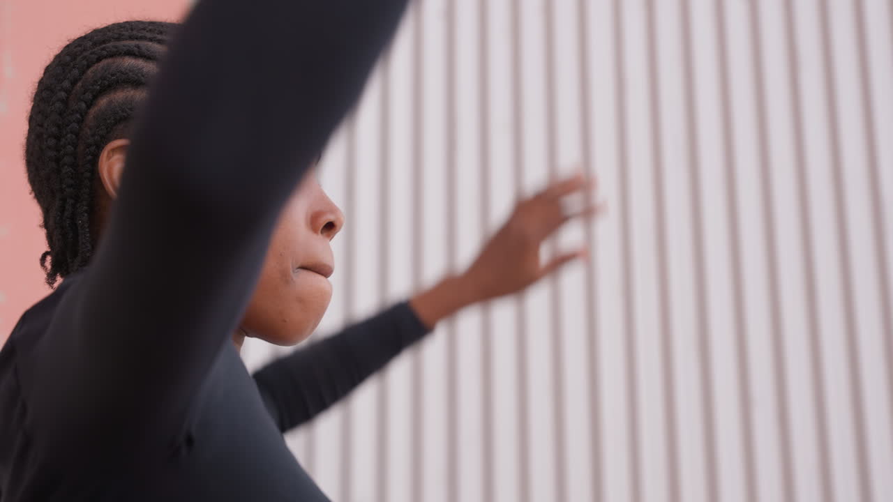 Giovane ballerina di colore prova movimenti controllati contro un muro con texture. Giovane ballerina di colore con capelli intrecciati esegue movimenti precisi delle braccia durante una sessione di pratica di danza urbana.