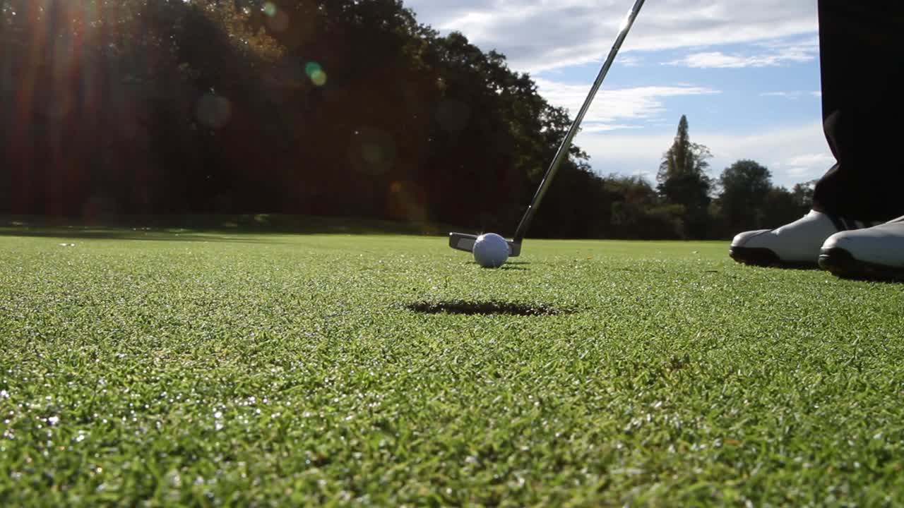 Ground level static shot of a golfer ptting into the hole, putting towards camera with lens flare.