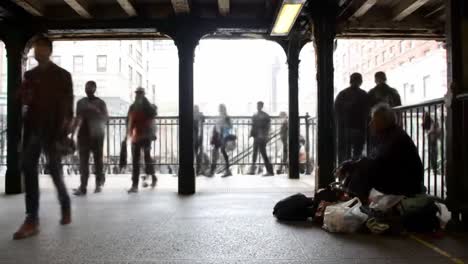 In a busy urban area, individuals hurry past a subway entrance while one person sits on the ground, surrounded by bags, amidst the ongoing activity of the city.
