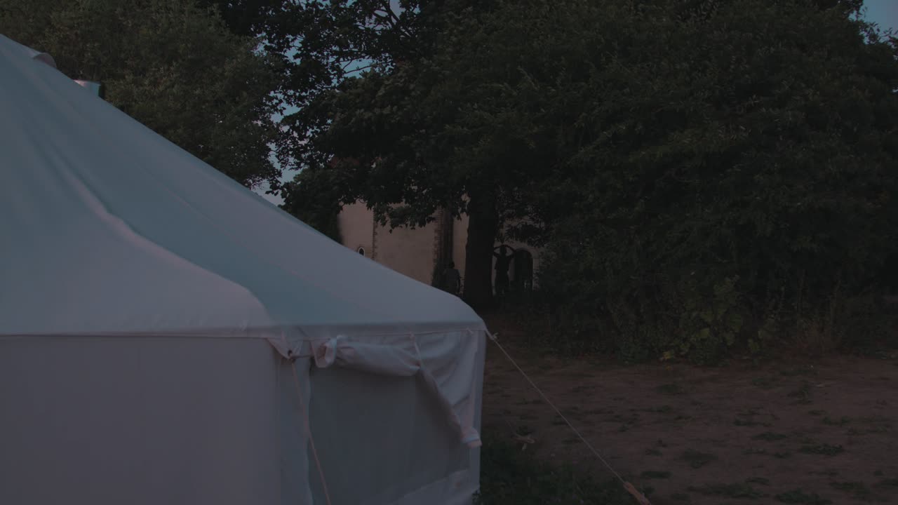 White Tent Set-Up At Foreground Of A Manor Home With Kids Playing Through Dusk. - Static Shot