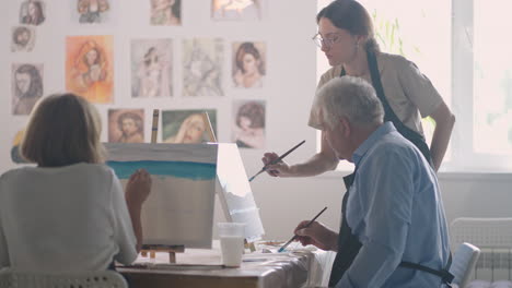 High-angle-view-of-cheerful-senior-friends-painting-on-canvas.-Senior-woman-smiling-while-drawing-with-the-group.-Seniors-Attending-Painting-Class-Together.-Senior-men-having-fun-painting-in-art-class