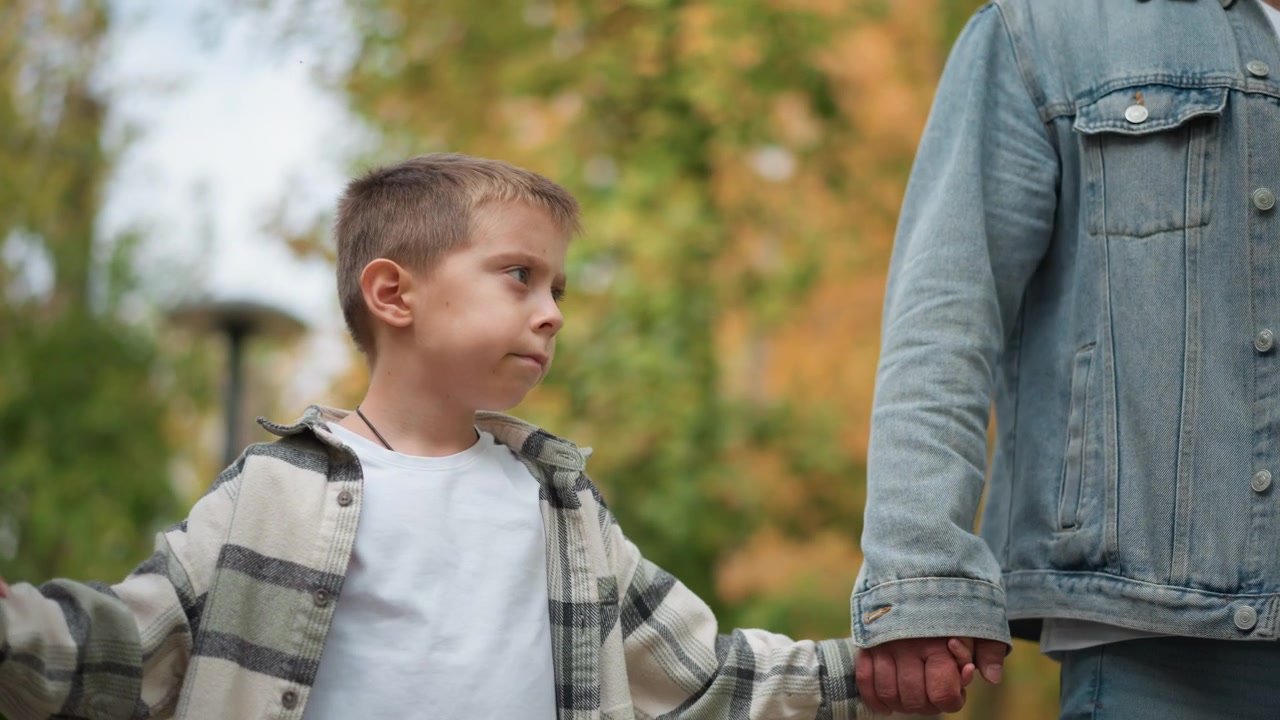 Primo piano di un bambino in piedi tra due adulti che gli tengono le mani, tutti all'aperto con vegetazione sullo sfondo. Il bambino indossa una camicia a quadri sopra una maglietta bianca, l'adulto a sinistra ha una giacca beige.