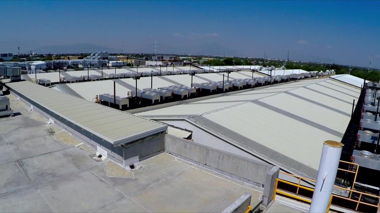Vista aerea con drone che mostra un grande complesso industriale con tetti in metallo bianchi, strutture di servizio e aree pavimentate aperte sotto un cielo azzurro e limpido durante il giorno in un'area commerciale.