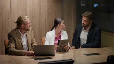 A-trio-of-office-workers-in-business-suits,-two-guys-and-a-girl,-discuss-solving-their-issues-using-a-laptop-and-an-electronic-tablet.-Business-meeting-of-office-workers-in-a-modern-office-with-wooden-flooring