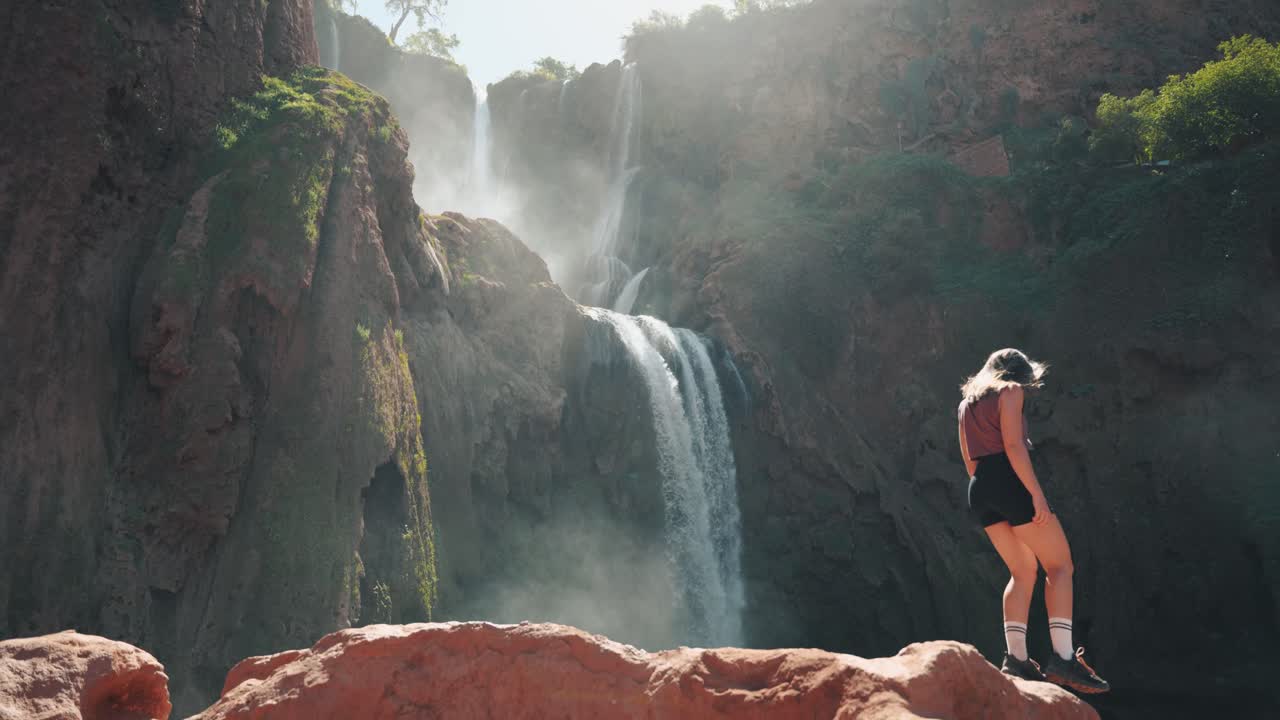 joven mujer caucásica caminando por las rocas frente a las cascadas en ouzoud, marruecos