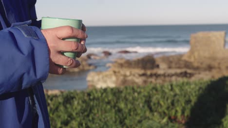 person holding cup of tea or coffee outdoors, ocean coast in background, copyspace for titles